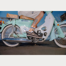 Load image into Gallery viewer, A person sitting on a light blue motorbike wearing hemp sneakers with a sunset design on the upper part.