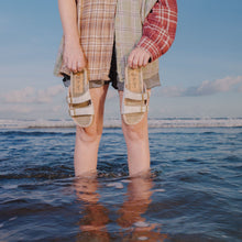 Load image into Gallery viewer, Person standing in water holding sandals with a beach and sky background